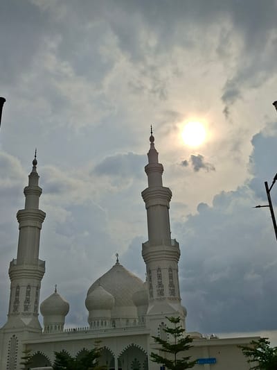 The image shows the facade of a white building with Islamic architecture, possibly a mosque, angled against a cloudy sky with the sun visible. The building features minarets, domes, and decorative latticework. The location is Ranto, Indonesia. There are trees partially visible on the right side of the frame. A light pole is visible in the upper central area of the picture.