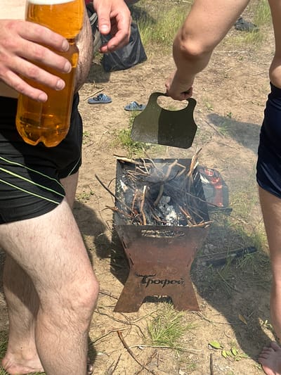 The image shows a group of people at an outdoor gathering, likely for a barbecue or camping trip, in Pravda, Russia. The scene is set on a sandy ground with patches of grass and dry twigs, suggesting a natural, possibly rural, environment. The time of day appears to be daytime, indicated by the strong sunlight casting shadows.

The main subjects are people, though their bodies are partially cropped. We see legs and arms, suggesting they are sitting or lounging. One person is holding a large plastic bottle of beer, and their hands are visible as they grip the bottle. Another person's hand is seen near a metal grill, holding a metal lid or plate. The grill itself is filled with burning wood and smoke, indicating a fire is actively burning.

In the background, a pair of blue flip-flops are visible on the ground, adding to the casual outdoor atmosphere. The people are dressed in casual clothing, such as dark shorts, further reinforcing the relaxed, informal setting.

The overall mood seems to be one of leisure and enjoyment, with the focus on a communal activity involving a campfire and drinks. There are no explicit emotional cues, but the activity itself suggests relaxation and socializing.

A notable detail is the visible text on the side of the metal grill, which reads "Гроссей" (Grossei) in stylized lettering. The weather appears to be sunny and warm, suitable for outdoor activities. The location context of Pravda, Russia, places the scene within a specific geographical and cultural setting.