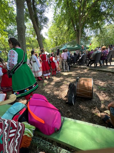This image captures an outdoor cultural gathering in Bolyarovo, Bulgaria, under bright daylight. The scene is set in a park-like environment characterized by tall trees, dense green foliage, and sun-dappled ground with sparse dirt and fallen leaves.
In the immediate foreground, a green wooden bench holds a vibrant pink backpack with an orange zipper accent, a light blue fanny pack, and a maroon textile with a white pattern. To the right, a large, light brown drum with white lacing is resting on the ground beside a black instrument case. A person's lower leg and foot, wearing dark sandals, are visible on the far right, suggesting they are seated nearby.
The mid-ground is populated by numerous individuals. Several women and children are dressed in traditional Bulgarian folk costumes, featuring white embroidered blouses, colorful aprons, and long skirts in shades of green, red, and white. One woman, prominent on the left, wears a green apron dress with floral embroidery and a white embroidered blouse, carrying a black shoulder bag. Other costumed individuals are gathered behind her, some facing the main activity.
Further back, a diverse audience, including many older adults, is seated on black plastic chairs, facing a green canopy tent. A man in a light blue polo shirt and sunglasses stands near the tent, observing the crowd. Colorful bunting flags are strung between the trees, adding to the festive atmosphere. The background consists of more trees, with sunlight filtering through the canopy.