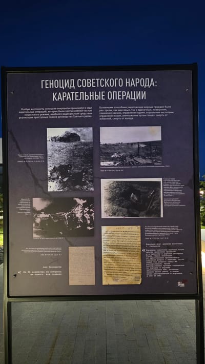The image shows a vertical display panel, likely an outdoor exhibit or informational sign, about the genocide of the Soviet people and punitive operations during wartime. The location is unspecified, but the panel's text is in Russian, and one caption refers to villages in the Smolensk region.
The panel features a dark gray background with lighter gray text and monochrome photographs. The text headings are "GENOCIDE OF THE SOVIET PEOPLE: PUNITIVE OPERATIONS." The body of the text describes the cruelty of German occupiers during punitive operations. It lists methods of destruction such as mass shootings, burnings alive, poisoning, starvation, torture, and freezing.
There are four black and white photographs arranged in a grid. These include:
* A picture of arsonists burning a village east of Smolensk.
* A picture of the village of Kozmodemyanovka, a picture of the burning expedition against the partisan territory.
* A close-up photo of a group of people examining a site where a chemical weapon was used, apparently in the Suzem district.
* A photo of a written document, presumably an official record.
Additional text includes captions for each photograph and quotes or excerpts from historical documents related to the atrocities committed. At the bottom of the panel, there is an “Act of Lawlessness”, and a statement indicating that “Of the 31 households, not a single one remained: all burned.”