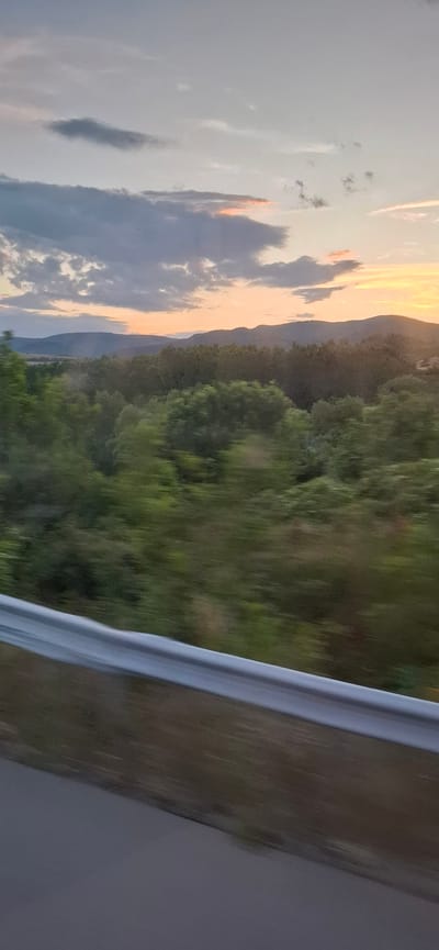 The image captures a scenic view of rolling hills and a dense forest, likely taken from a moving vehicle on a road. In the foreground, a silver guardrail runs horizontally across the frame, indicating the presence of a highway or a similar road. The lush green trees of the forest dominate the mid-ground, showing motion blur consistent with a fast-moving vehicle. Beyond the forest, a series of mountains or hills are visible, silhouetted against a sky filled with a mixture of grey and pale orange clouds, suggesting either dawn or dusk. The sky exhibits a gradient of colors, with warmer tones towards the horizon where the sun is setting or rising. The scene appears to be a natural landscape, with no visible buildings or signs of urban development other than the road infrastructure. Given the query, the location is the region of Djerman, Bulgaria, an area known for its natural beauty and mountainous terrain, which aligns with the visual elements in the photograph.