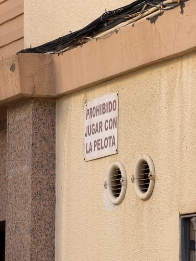 The image shows a close-up, low-angle view of a building exterior, likely a wall or ceiling, in an urban setting. The dominant textures are a rough, stucco-like beige wall and a section of speckled granite. Two round ventilation grates are visible on the beige wall, set at an angle. To the left, a thick bundle of black electrical wires is taped to a beige structural beam that also has a rough texture. The beam appears to be part of an archway or overhang, with cracked paint and plaster.
Attached to the beige wall, between the ventilation grates, is a rectangular white sign with black text. The sign is slightly tilted and mounted with visible fasteners. The text on the sign reads: "PROHIBIDO JUGAR CON LA PELOTA". This translates from Spanish to "PROHIBITED TO PLAY WITH THE BALL". This indicates a rule or restriction in the area.
Below the beige wall and granite section, a dark horizontal element is partially visible at the bottom right, possibly the top of a doorway or another architectural feature. The lighting suggests it is daytime, with soft, diffused light, and there are no clear indicators of the weather. The overall scene is utilitarian and slightly weathered, common in many built environments. The focus is on the architectural details and the warning sign.