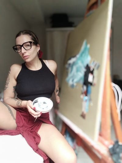 The image depicts a woman with tattoos, glasses, and a black tank top, seated and holding a paint palette with a brush, positioned in front of a large canvas on an easel. The painting on the canvas is partially visible. The woman wears a red skirt. The location appears to be an indoor studio space. There is no verifiable evidence to confirm that the location is in Inriville, Argentina.