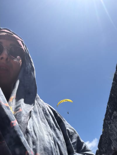 A low-angle, slightly tilted shot features a person wearing sunglasses, with a patterned fabric covering their head and shoulders. A yellow paraglider is visible in the blue sky, and a section of a stone structure is at the top of the frame. The location is inferred to be Benoa, Indonesia.