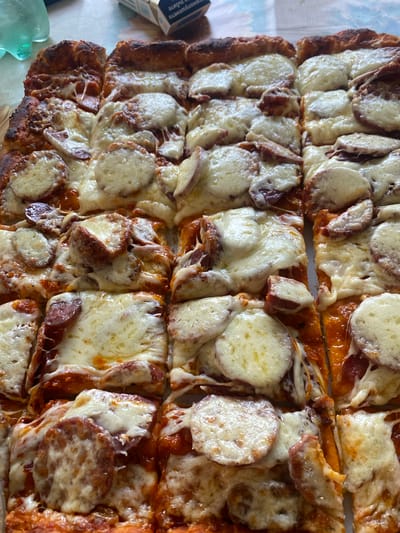 A close-up overhead shot captures a rectangular pizza, cut into numerous slices, presented on a surface that appears to be a patterned tablecloth. The pizza is generously topped with melted cheese and slices of pepperoni, some of which have crisped edges. The crust shows a well-baked, slightly charred texture. In the background, out of focus, a green plastic bottle and a pack of cigarettes are visible, hinting at a casual setting, possibly a picnic or an outdoor meal. The lighting is natural, casting a soft glow on the food. The scene is devoid of any human presence or activity. The overall impression is one of a delicious, ready-to-eat meal.