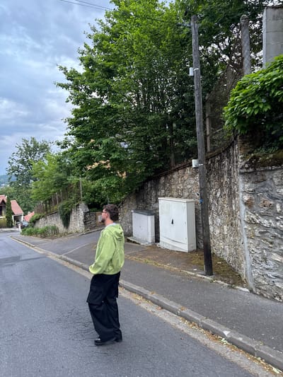 The image displays an outdoor street scene in Orsay, France, on an overcast day. A young man stands on an asphalt road, facing towards the left side of the frame, next to a weathered stone wall. He is wearing a light green long-sleeved shirt, black wide-leg trousers, and dark shoes. He has short dark hair, a beard, and glasses, and appears to be looking downwards or to his immediate left.

To the man's right, along the stone wall, a wooden utility pole stands tall. Attached to the pole is a small white device. Below it, at the base of the stone wall, two grey and white utility or electrical boxes are mounted on a concrete slab. The stone wall itself is rugged, built from various sizes of light-colored stones, and shows patches of moss and greenery. Behind the utility pole and the wall, a dense mass of green foliage and trees is visible, with a chain-link fence partially obscured by the vegetation.

The asphalt road features a distinct curb bordering a narrow dirt strip next to the stone wall. The sky above is entirely covered with grey clouds, indicating an overcast or heavily cloudy day, with no direct sunlight. In the distance, beyond the trees, hints of residential buildings with red-tiled roofs can be seen nestled on a hillside. There is no discernible text visible on any objects in the image.