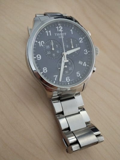 A Tissot chronograph wristwatch, predominantly silver-toned and metallic, is depicted resting at an oblique angle on a light-colored wooden surface. The scene is located in Fontenay-sous-Bois, France.

The watch features a round case and a three-link metal bracelet, both exhibiting a polished finish. Its dark grey dial is marked with white Arabic numerals from 1 to 12 and includes three sub-dials. A date window is positioned between the 4 and 5 o'clock markers, displaying the numeral '9'. The brand name "TISSOT" and "1853" are printed below the 12 o'clock position, with "SWISS MADE" visible near the 6 o'clock mark.

The white hour hand points approximately to 7:55, and the minute hand points to approximately 10:55. The central thin hand, a chronograph second hand, is reset to the 0/60 position. The small hand in the bottom sub-dial, the running seconds counter, indicates approximately 20 seconds. A fluted crown and two pushers are visible on the right side of the watch case.

The background consists of a uniformly lit wooden surface, displaying subtle grain patterns. No individuals or active movements are present within the frame.