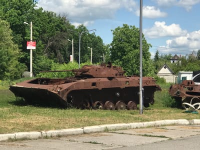 Two heavily rusted military vehicles, likely infantry fighting vehicles (BMPs), are prominently displayed outdoors on a grassy area. The vehicle in the foreground is largely intact but severely corroded, with its gun barrel pointing towards the left. Its tracks are partially missing, and the interior appears empty and exposed through open hatches. To the right, a portion of a second similar, equally rusted vehicle is visible, also in a state of disrepair.
The vehicles rest on a patch of green and dry grass, bordered by a cracked concrete curb and a paved surface in the foreground. A lush backdrop of dense green trees dominates the middle ground and background, suggesting a natural or semi-urban setting. Above the trees, a bright blue sky with scattered white cumulus clouds is visible.
Several tall light poles with streetlights are positioned throughout the scene. One pole stands centrally in front of the main vehicle. In the distance, partially obscured by trees, residential buildings with distinct peaked roofs can be seen.
A red sign with white Cyrillic text is attached to a light pole on the far left. The text is legible as "ЗАПРОШУЄМО на роботу" (We invite to work), followed by a phone number: "067 2 333 777". The scene depicts a bright, clear day, indicated by the lighting and cloud cover. The extreme rust and derelict condition of the vehicles suggest they are abandoned, decommissioned, or perhaps serve as a static display or monument.
