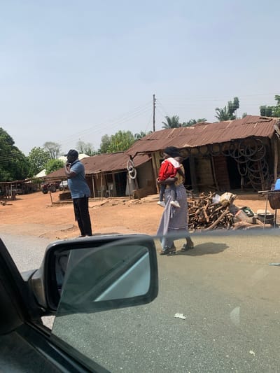 The image depicts a street scene in Karshi, Nigeria. A woman wearing a red shirt and carrying a child stands in front of a corrugated metal-roofed building, possibly a shop. The building has tires and firewood stacked near its entrance. A man in a blue shirt and black pants stands nearby, appearing to be using a phone. The scene is viewed from inside a vehicle, with the side mirror visible in the foreground. The street is paved and adjacent to a dirt area with other structures and vegetation visible in the background under a clear sky.
