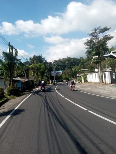 A daytime image depicts a curved asphalt road in the city of Ban Phonhxavanh, Laos, under a bright blue sky with scattered white clouds. Long, dark shadows, likely from overhead power lines, stretch across the road surface. On the left side of the road, a person wearing a blue jacket and a white helmet operates a dark motorcycle, traveling away from the camera. Another person on a red motorcycle, without a visible helmet, is traveling in the opposite direction on the right side of the road. Multiple other motorcycles and at least one light-colored car are visible in the distance. The road is marked with solid white lines along its edges. To the left, a utility pole heavily overgrown with vines supports power lines, adjacent to small structures with corrugated roofs and dense green foliage. To the right, a white-walled structure, a dark gate, and tall trees line the roadside. In the background, a large billboard partially displaying "THERMAL RESORT" text is situated before a densely forested hillside dotted with residential buildings.