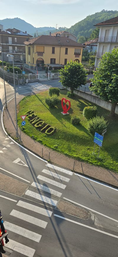 The aerial image depicts a bright, sunny day in Cengio, Italy. The scene is dominated by a green roundabout at a road intersection. On the roundabout, the word "CENGIO" is spelled out in sculpted hedges. A bright red, abstract heart sculpture stands next to the hedge lettering. Road signs indicating directions to "COSERIA 4", "TORINO SAVONA", "MILLIESIMO", and "SAVONA 33" are visible on the right side of the roundabout. Across the street, a pedestrian crossing with white stripes is marked on the asphalt. Several residential buildings with tiled roofs and balconies are visible in the background, nestled against lush green hills. The overall impression is a peaceful, well-maintained urban landscape.