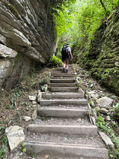 A person with a backpack walks up a stone staircase in a narrow, forested gorge. The staircase is made of concrete slabs and is flanked by moss-covered rock walls on either side. Lush green vegetation grows on the walls and along the sides of the path. The lighting suggests it is daytime, possibly overcast. This scene appears to be a natural attraction or a hiking trail, potentially within the city of Khleborob, Russia, given the prompt's context.