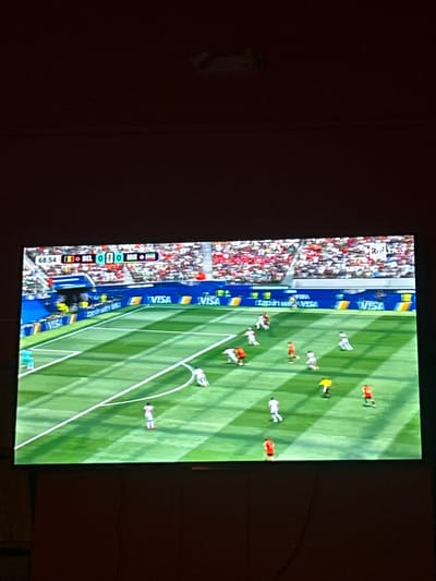 The image shows a television screen displaying a soccer match. The score displayed is 0-0 between Belgium (BEL) and Iran (IRN) at 68:54 minutes. Several players in white and red uniforms are visible on the green field, which has white lines marking the boundaries and penalty areas. The stadium appears to be full of spectators wearing red clothing. Advertising banners, including "VISA," are visible along the edge of the field. The top right corner of the screen shows "RAI LAD." The television is placed against a dark brown wall. The scene is viewed from a slightly elevated angle. The location in Militello in Val di Catania, Italy, is not visually represented in the image itself; it is inferred context.