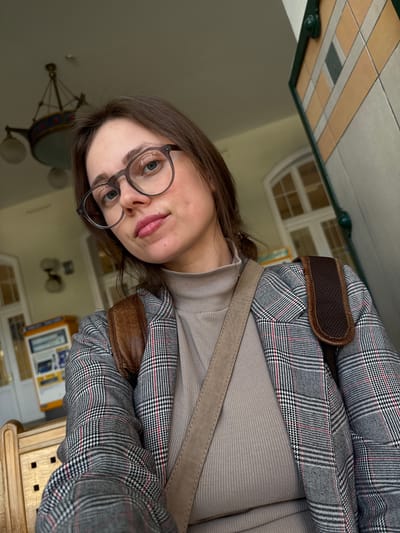 A young woman with light brown hair, blue eyes, and fair skin is captured in a close-up, slightly upward-angled selfie. She wears round, dark-framed glasses and has a playful expression, with her tongue subtly peeking out. She is dressed in a light beige or tan ribbed turtleneck top, overlaid with a stylish grey and white plaid blazer featuring thin red accent lines. A brown, possibly leather, strap from a bag, with a padded shoulder section, crosses her body.
The setting is an indoor public space, possibly in Szeged, Hungary. The background shows a distinct ceiling composed of beige and grey rectangular tiles. A decorative chandelier or light fixture with a circular base and multiple arms hangs from the ceiling. A window with multiple panes, likely arched or rounded at the top, is visible, suggesting a traditional architectural style. Other blurred elements in the background, including what appears to be a wooden counter or furniture in the lower right, indicate a bustling interior environment, perhaps a waiting area, station, or hall during the daytime. No discernable text is visible.