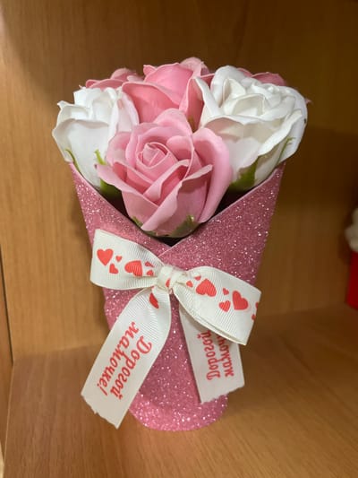 A bouquet of artificial pink and white roses is displayed in a pink, glitter-covered conical vase. A white ribbon with red hearts is tied around the vase, featuring text in Cyrillic script that translates to "To my dear little one!". The background consists of a wooden surface, likely a shelf or table. The image provides no information about the location in Pokoinoe, Russia.