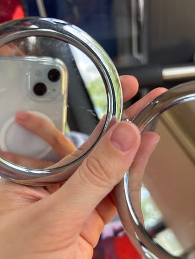 The image shows a close-up of a person's hand holding a round, metallic compact mirror. The mirror is open, and its reflective surface shows a distorted reflection of the person's finger, a smartphone with two camera lenses, and a white circular object on the back of the phone. The background of the reflection is blurred, showing hints of green foliage and a bright, out-of-focus light source, suggesting an outdoor setting, possibly during the daytime. The hand holding the mirror appears to be lightly tanned and has short, clean fingernails. The mirror itself has a polished silver-toned frame. The scene is intimate and personal, focusing on the act of holding and using a personal grooming item. The lighting suggests it's a bright day. The context "Dolní Vinice, Czechia" implies the location where this photograph was taken.