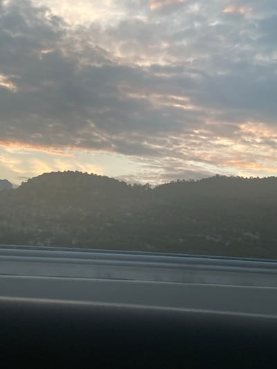 This media file displays a landscape scene dominated by a cloudy sky and a tree-covered mountain range. The clouds are textured and appear to be catching the light of either sunrise or sunset, with hints of orange and yellow interspersed with grays. The mountain range forms a dark silhouette against the sky, its edge clearly defined by the light. To the right of the frame, a portion of what appears to be a window or door frame is visible, with a metallic or plastic finish that suggests an indoor perspective looking out. The overall mood is serene and atmospheric, capturing the beauty of a natural landscape bathed in soft light. The time of day is likely dawn or dusk due to the lighting conditions on the clouds and the silhouette of the mountains. No people, objects, or visible text are present in the image. The location context is Donje Mrke, Montenegro.