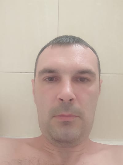 The image is a close-up, head-and-shoulders photograph of a light-skinned man with short, dark hair, a receding hairline, and a light stubble on his face. He has dark brown eyes and a neutral expression. The man is shirtless, revealing his shoulders and chest with some dark hair. The background consists of a light-colored, tiled wall. The lighting appears to be artificial and is slightly warm. There are no discernible landmarks or features that would indicate a specific location within the city of Kobeliaky, Ukraine. The photograph is taken from a frontal perspective, with the man's face filling most of the frame.