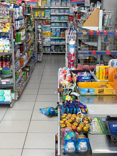The image depicts the interior of a convenience store, likely a minimarket, in Denasri Kulon, Indonesia. The perspective is from a slightly elevated position looking down an aisle. The store is well-stocked with various consumer goods displayed on shelves and racks.

On the left side of the frame, shelves are filled with packaged products, including what appear to be toiletries, beverages, and food items. Notable products visible are bottles of dark liquid, possibly sauces or drinks, and boxes of cereal. Further down the aisle on the left, there are stacks of packaged facial tissues.

The central and right portions of the image showcase more shelves and displays. These shelves are laden with a variety of items, including diapers, baby products, and packaged snacks. A prominent display features numerous colorful lollipops and candies arranged on a counter or checkout area. Among these sweets are Kinder Joy eggs. Also visible on this counter are payment terminals, suggesting this is a transactional area. A blue bag of chips is on the floor in the middle of the aisle.

The flooring is tiled with light-colored squares. The lighting appears to be artificial, typical of an indoor retail environment. No individuals are visible in the image. The overall scene is one of a clean, organized retail space, characteristic of minimarkets in Indonesia.
