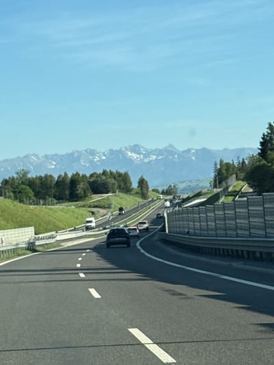 The image shows a highway curving through a mountainous landscape under a clear blue sky. The road has multiple lanes with markings, and several vehicles are visible. A sound barrier runs alongside the highway in sections. The mountains in the background appear to be snow-capped. The location of the scene is not apparent from the image. Therefore, the city of Klikuszowa, Poland is not indicated in the image.