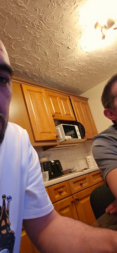 This rotated image offers a unique, slightly tilted, overhead perspective of an indoor domestic scene, likely a kitchen or dining area in Norwich, United Kingdom.
At the very top of the frame, a person is visible upside down, appearing to be taking a selfie. Only a portion of their face, including eyes, nose, and a beard, is visible, along with the shoulder and arm of a white t-shirt. The t-shirt features a distinctive graphic design in black, gold, and blue, which resembles a stylized crown or an insect with antennae.
In the lower part of the image, a second individual is partially seen, wearing a grey t-shirt. Only their head, shoulder, and arm are visible, and they appear to be looking downwards or away.
The room is characterized by extensive wooden cabinetry with a warm, light-brown finish and traditional paneling, fitted with small, round brass or gold-colored knobs. A white microwave with a black glass front is built into one of these cabinets on the right side of the frame. Adjacent to it, a black kitchen appliance, possibly a coffee machine or toaster, rests on a light-colored countertop. The floor is covered with light, square tiles.
The wall on the right side of the image features a pronounced, decorative texture with swirling or feather-like patterns. A bright, warm light source, likely a light fixture, illuminates this textured wall from the bottom right corner. The scene depicts a casual, candid moment within a home setting, bathed in artificial light. No visible text is present in the image.