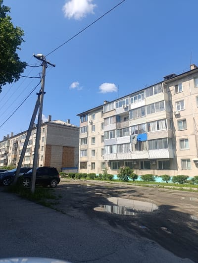 The image captures a street scene in Svobodny, Russia, on a bright, clear day. Two apartment buildings dominate the right side of the frame, their concrete facades weathered and showing signs of age. The building closer to the foreground has multiple balconies, many enclosed with glass, and several windows. Air conditioning units are visible on some of the walls. The building further back appears to be constructed of light-colored brick with darker brick accents. A utility pole with multiple wires stretches across the upper left portion of the image, with a streetlamp attached to it. In front of the buildings, a row of green bushes lines a turquoise-painted curb. The street itself is paved with asphalt and features several large puddles, indicating recent rain or an issue with drainage. Two cars are parked on the left side of the street: a dark-colored SUV and another dark vehicle behind it. Trees with green foliage are visible on the far left. The sky is a vibrant blue with a few scattered white clouds.