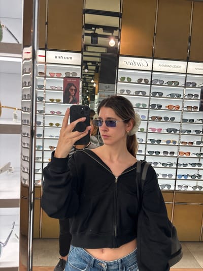 A woman in sunglasses and a black zip-up jacket takes a mirror selfie in what appears to be an eyewear store. Rows of sunglasses are displayed behind her, categorized by brand, including Cartier and Dolce & Gabbana. She is holding a black phone with a pop socket in her hand. She also wears low-rise jeans. Her reflection reveals store details, including lighting fixtures and more eyewear displays.