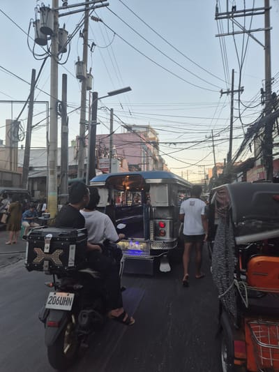 The image depicts a street scene in Pasay City, Philippines. Overcrowded utility poles with a dense network of wires line the street. A variety of motorized tricycles, known locally as "tricycles" or "motorized pedicabs," are parked or moving along the road. The tricycles exhibit diverse designs and colors. Several people are visible, some walking or standing near the vehicles, while others are riding. A motorcycle with a rear storage box and license plate number 206 UHN is also visible. The environment appears to be a densely populated urban area with buildings and storefronts lining the street.