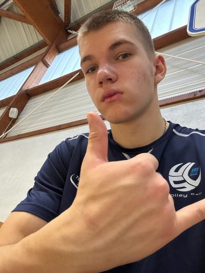 A young adult male, with short light brown hair and light eyes, is depicted in a close-up, chest-up selfie format. He maintains a neutral expression with slightly pursed lips, looking directly into the camera. He wears a dark blue short-sleeved athletic shirt with white trim. On his right chest (viewer's left), a white logo is visible, featuring a stylized volleyball design, the letters "SUC," and the partially legible text "Volley Ball." A silver chain is around his neck. His right hand, prominent in the foreground, is positioned to display a "shaka" sign (thumb and pinky extended, middle fingers folded). The background indicates an indoor sports facility, likely a gymnasium, with exposed wooden roof beams, white corrugated ceiling panels, a white wall-mounted speaker, and a metal grate structure hanging from the ceiling. This scene is located in Sennecey-le-Grand, France.
