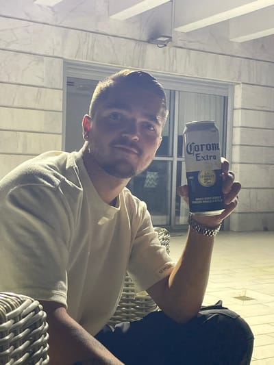 A male individual is depicted seated at night on what appears to be an outdoor patio or balcony in Yavne, Israel. He is a young adult with short dark hair, light facial hair, and an earring in his left ear. He wears a light-colored, possibly cream or off-white, ribbed short-sleeved shirt with a small label reading "HERMANO" on the left sleeve. His right hand is raised, holding an open silver-colored can of Corona Extra beer, while a metallic link bracelet or watch is visible on his right wrist. Partially visible wicker-style furniture is present in the foreground. The background features a building facade constructed of light-colored, possibly marble or stone tiles, with a glass sliding door or window. The scene is illuminated by an overhead artificial light source, casting subtle shadows. The ground surface is a light-colored tiled patio.