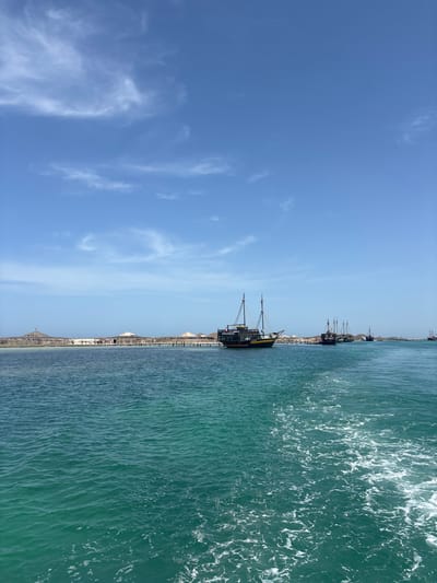 The image captures a view from a boat on the sea, looking towards a coastline. The water in the foreground is a vibrant turquoise, with white foam trailing from the moving vessel. In the distance, a sandy shoreline is visible, lined with thatched-roof umbrellas and structures, suggesting a beach resort or leisure area. Several traditional-looking wooden boats, some resembling pirate ships, are anchored or cruising in the water near the shore. The sky above is a clear, bright blue with wispy white clouds. This scene is consistent with the coastal areas of Gizen, Tunisia, known for its beautiful beaches and maritime activities.