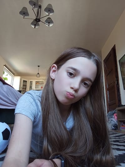The image is a selfie of a young girl with long, brown hair, taken indoors. She is wearing a grey t-shirt. Her hair is prominent, covering a significant portion of the frame. Her face is tilted, and she has her lips slightly pursed. She appears to be looking towards the camera with a neutral or slightly playful expression.
In the background, various objects and parts of a room are visible. To the left, there's a glimpse of a black and white stuffed soccer ball. Further back, a light-colored cabinet with glass doors is visible, possibly containing books or decorative items. A skylight is open at the top, suggesting it might be daytime. A modern, multi-headed ceiling lamp with a metallic finish is mounted on the wall on the right side of the image. Below the girl's hair, a wooden door and a shelf with some items, including plastic bags and what appear to be art supplies, can be seen.
The overall setting suggests a casual indoor environment, likely a home. The lighting appears to be natural, possibly enhanced by indoor lights, and there are no immediate indicators of the specific time of day beyond the skylight. The image is captured in portrait orientation, with the subjects and scene rotated. No visible text is present in the image.