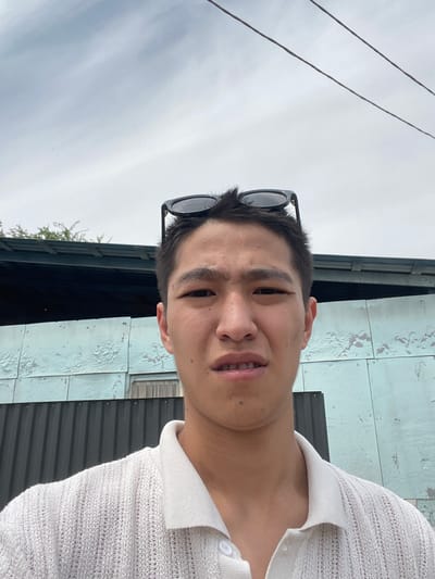 The image is a waist-up, selfie-style photograph of a young East Asian male taken outdoors in Кенен Әзірбаев ауылы, Kazakhstan. The man is wearing a white, short-sleeved, collared knitted shirt. His black sunglasses are resting on top of his head. He has short, dark hair and is looking directly at the camera with a slightly contorted facial expression, his mouth open. In the background, there is a pale blue, weathered building with visible paneling, and above that, a dark roofline and some green foliage. Two power lines are visible against the cloudy sky. A dark corrugated metal fence is positioned in front of the building. The lighting suggests it is daytime, likely overcast.