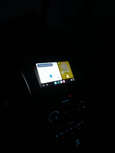 The image depicts the interior of a vehicle at night or in low-light conditions, specifically the dashboard and central console. The location is Vinnytski Khutory, Ukraine. A central infotainment screen is illuminated, displaying a split view of two applications. The left side shows a navigation interface with a map featuring a blue vehicle icon and Ukrainian text "Переглянути оновлення" (View updates). The right side shows a music player interface, displaying album art and the song title "Touch Too Much" by "AC/DC". Below these applications, a row of app icons includes a microphone, Google Assistant, and Spotify. The time "17:05" is visible in the bottom right corner of the screen. Below the infotainment screen, the climate control panel and other vehicle controls are visible, illuminated with a blue backlight. Elements include a "PASSENGER AIRBAG" indicator, "VOL" (Volume) and "TUNE" rotary dials, and various push-buttons for climate control functions such as fan speed, temperature adjustment, "AUTO," "A/C," "MAX A/C," and heated seats. No occupants are visible within the frame.