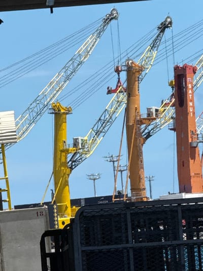 The image displays a detailed view of the Port of Papeete, France, under a clear blue sky. The foreground is partially obscured by a dark, perforated metal structure and a grey panel marked '18'. The mid-ground and background feature multiple large port cranes and shipping containers. A prominent yellow crane with a lattice boom rises on the left. Central to the image is an orange-yellow gantry crane, marked '2' and 'LIEBHERR', with a red container spreader bar suspended from its hoist, positioned above dark blue shipping containers. One container is labeled 'CMACGM'. To the right, a reddish-orange crane, identified by 'SATNUI' and 'TEREX GOTTWALD', is partially visible. The containers are stacked on what appears to be a cargo vessel or the port quay. In the distance, several industrial floodlight masts are visible. The scene is brightly illuminated by daylight; no human figures are discernible.
