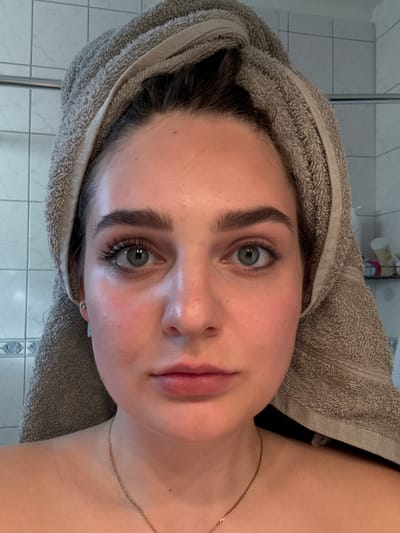 A close-up, eye-level portrait shows a young woman looking directly at the camera. Her head is tilted slightly to the right, and her dark hair is wrapped in a light brown, textured terrycloth towel, suggesting she may have just showered or is preparing for a skincare routine. She has fair skin, prominent dark eyebrows, and striking green or hazel eyes with visible eyelashes, possibly enhanced with mascara. Her lips are naturally pink and full. A delicate gold-colored chain necklace rests on her collarbone and shoulder.
The background, largely out of focus, reveals a tiled wall, likely in a bathroom. The tiles are light-colored, possibly white or light grey, with visible grout lines. On the upper left, a reflective metal bar or frame (possibly part of a shower enclosure) is visible. In the upper right corner, several blurred bottles or containers are visible on a shelf. The lighting is soft and even, illuminating her face clearly. The woman's expression is neutral and calm. There is no visible text in the image.