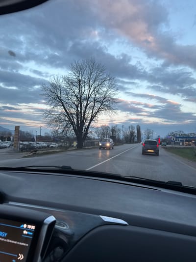The image depicts a roadside scene at dusk or early evening, observed from the interior of a vehicle through its windshield. The location is Piperkov Chiflik, Bulgaria.

The foreground shows the dark gray dashboard of the observation vehicle, with a portion of a digital infotainment screen visible on the lower left displaying "18:35" and a signal strength icon. Windshield wipers are present at the base of the windshield.

The main subject is a two-lane asphalt road, which appears wet, indicating recent precipitation. Two vehicles are visible on the road: a dark gray hatchback (license plate "EB 9624 KE" partially visible) is traveling away from the camera in the same direction, and a light-colored hatchback with headlights illuminated is approaching in the opposite lane.

To the left of the road, a large, bare deciduous tree stands prominently. Behind it, a car dealership or lot contains numerous parked vehicles. Further left and partially obscured, a multi-story structure is visible. Distant mountains are present on the horizon under a cloudy, blue-gray sky streaked with pink and orange hues from the setting sun.

To the right of the road, a Petrol-branded gas station with fuel pumps and a service building is visible. Digital signage displaying red numbers (possibly fuel prices) is present. Utility poles and power lines line the roadside, along with additional bare trees further down the road. A third light blue vehicle is partially visible near the gas station. No individuals are explicitly discernible in the scene.