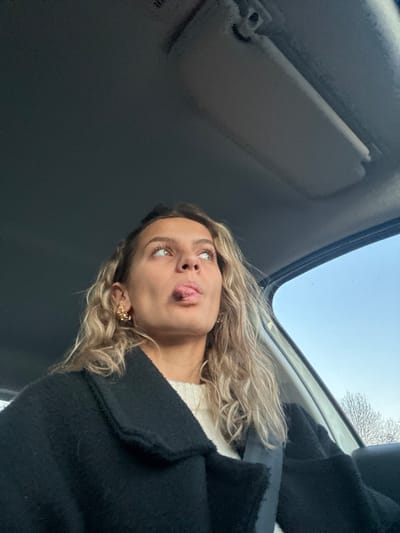 A woman is photographed from below inside a vehicle. She sticks her tongue out, her eyes are wide, and she is wearing a black coat over a white sweater. The sun visor is visible, and the sky is seen through the sunroof. Her blonde hair is loose and wavy, and she wears ornate gold earrings.