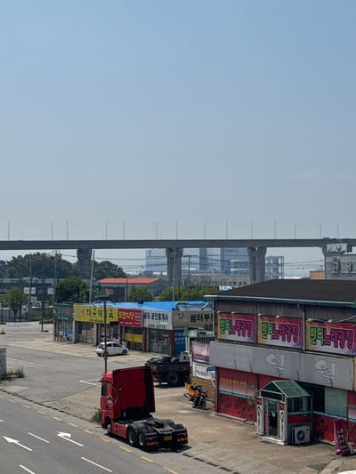 The image shows a street scene in Pyeongtaek-si, South Korea, with a large overpass dominating the background. In the foreground, a red semi-truck is parked on the side of a road with white directional arrows painted on the asphalt. To the right of the truck, a commercial building with several signs is visible. One sign, in Korean, has the text "반도노래광장" repeated three times in colorful lettering. Another sign, also in Korean, reads "엄청노래방". A small green glass-covered entrance booth is situated in front of the building, with an air conditioning unit mounted below it. A motorcycle is parked near the building to the left. Further down the street, several smaller shops with awnings are present, and a white car is parked in front of one of them. The sky is clear and blue. The overall scene depicts a commercial area with transportation infrastructure.