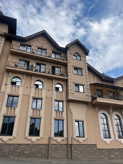 The image captures a detailed, tilted view of a multi-story building against a partly cloudy sky. The architecture is elaborate, featuring a light brown or beige facade with dark brown accents on the roofs, window frames, and decorative elements.
The building incorporates various window designs, including rectangular, semi-circular, and arched shapes, many adorned with decorative surrounds and cornices. Multiple balconies with intricate, ornate black metal railings are visible, adding to the building's sophisticated aesthetic. One prominent gable on the upper left features a circular, quatrefoil-like decorative motif. Sections of the exterior wall, particularly on the right side, are finished with a contrasting grey, textured stone-like material.
An air conditioning unit can be seen mounted below one of the balconies. Drainage pipes run down the building's exterior. The sky is blue with scattered white clouds, indicating daytime and fair weather. The overall style suggests an eclectic or possibly neo-Gothic architectural influence. No human activity or visible text is present in the image. The context for the location is Kamianytsia, Ukraine.