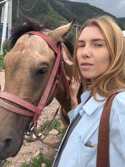 The image shows a close-up selfie of a young woman with blonde hair leaning against a horse's head. The woman is wearing a light blue collared shirt with a brown strap over her shoulder. She has a relaxed expression. The horse has a light brown coat and is wearing a red bridle. The background features a lush, green mountainous landscape with trees, suggesting an outdoor setting. The sky is overcast, indicating a cloudy day. The terrain appears to be rocky and grassy. The woman's left hand is resting on the horse's neck. There is a visible blue embroidered symbol, possibly a "20," on the woman's shirt pocket. The scene is likely taking place in Kaindy, Kyrgyzstan, given the mountainous and forested environment. The overall mood is peaceful and suggests an outdoor activity like horseback riding.