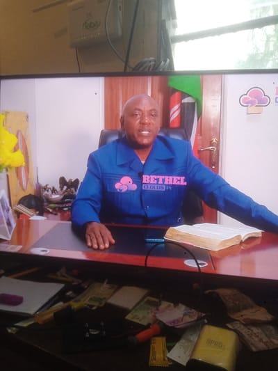 A television screen displays a man in a blue jacket with "BETHEL CLOUDS TV" visible, sitting behind a desk with an open book, likely a bible. A Kenyan flag is partially visible in the background. In front of the screen, a cluttered surface contains various items, including money, tools, and packaging. A ZTE router is attached to the wall near the top of the image.