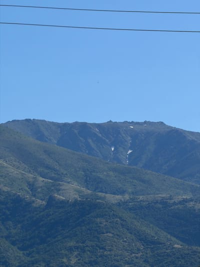 The image depicts a vast, mountainous landscape under a clear blue sky. In the foreground, there are several layers of hills covered in dense green vegetation. These hills create a textured, undulating terrain. In the middle ground and background, a larger, rugged mountain range rises. The upper slopes of this mountain are rocky and bare, with patches of snow visible in some crevices. The sky is a uniform, light blue, devoid of clouds. Three thin, dark horizontal lines, likely power lines, stretch across the upper portion of the image. The overall scene is one of natural grandeur and remoteness, characteristic of the Rila Mountains in Bulgaria.