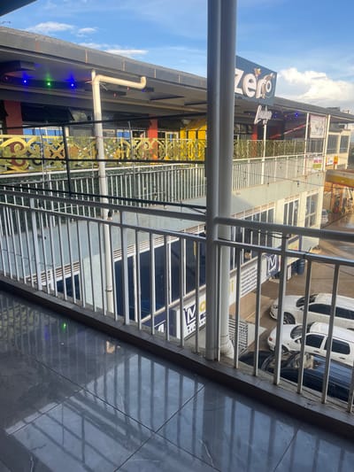 The image depicts an exterior view from an elevated position, likely a balcony, overlooking a multi-story commercial complex in Biashara ward, Kenya, under daylight conditions with a blue sky and scattered white clouds.

In the immediate foreground, a dark grey tiled floor reflects vertical white metal railing bars, which form a protective barrier. Directly below, an intermediate level features white metal railings and a green artificial hedge or covering on a balustrade.

Further down, the ground level or lower floor of the complex contains storefronts with large reflective windows. A visible sign on this level reads "MO" (partially visible) and "SERVING F...", with an adjacent list of categories: "LIQUOR STORE", "BAKING & COFFEE", "HOT AND COLD DRINKS", "BEVERAGES", "HOUSEHOLD GOODS". A prominent white structural pillar extends vertically through the scene. No individuals are visible on these lower levels.

The upper levels of the building feature a large sign with the word "zero" and a target-like graphic, accompanied by "Lounge" underneath, indicating a commercial establishment. This level is adorned with a gold-colored, ornate metal railing with scrollwork. Blue, green, and red LED lights are visible under the eaves of the upper floor structure.

To the far right, a yellow canopy with the red Shell logo indicates a gas station. A parking area below contains several parked white vehicles (sedans and SUVs) and one black vehicle. No people are discernible engaging in specific actions or interactions within the frame.