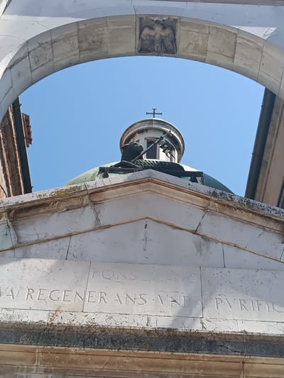 A low-angle shot of a building, possibly a church or chapel, framed by an arched doorway, most likely located in Mezzana Sant'Antonio, Italy. The building is constructed of white stone, with Latin inscriptions carved into its façade that read "FONS VA REGENERANS VNDA PVRIFIC." Atop the building is a green dome, and a smaller, circular structure with a cross on top. A statue of a lion with a banner is placed at the top of the building. The blue sky is visible through the archway and between the dome and the edges of the doorway. The doorway itself is also made of white stone, with a decorative carving of an eagle holding a sheaf of wheat at the arch's apex. Portions of adjacent buildings are visible on either side of the structure.