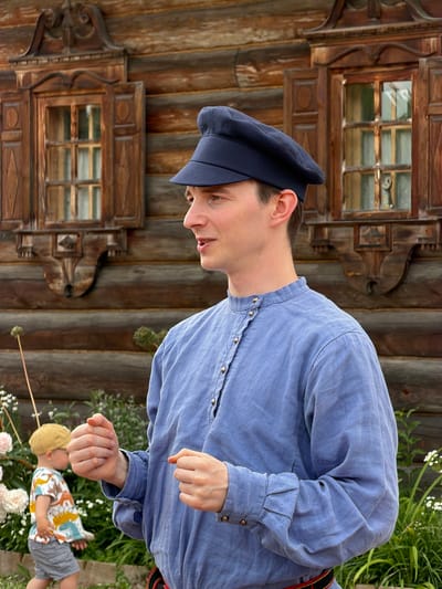 The image shows a man wearing a blue collared shirt and a dark blue cap, standing in front of a wooden house. The house has intricate carvings around the windows. In the background, a young child wearing a yellow cap and a colorful t-shirt is visible. The scene appears to be outdoors, with greenery and flowers present. The man is looking to the side and has his hands raised as if speaking or gesturing. The overall setting suggests a traditional or historical location, possibly in the city of Dnp Ryzhiki, Russia.