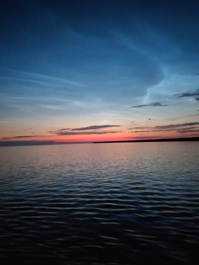 The image depicts a tranquil natural scene during sunset. The primary subjects are a vast expanse of calm water, reflecting the colors of the sky, and the sky itself, transitioning from deep blue at the top to vibrant hues of orange and pink closer to the horizon. Wispy clouds are scattered across the sky, catching the last light of the day.
The scene is set at dusk, with the sun having just dipped below the horizon, creating a beautiful gradient of colors. The water is smooth with gentle ripples, suggesting a peaceful and quiet environment. In the distance, a dark, silhouetted landmass is visible, marking the edge of the body of water.
The overall mood conveyed by the image is serene and contemplative, evoked by the soft light, the gentle movement of the water, and the expansive horizon. There are no people or specific objects visible, emphasizing the natural beauty of the landscape. The location cues suggest this is a body of water, possibly a lake or a sea, with land on the far side. The time of day is clearly sunset or twilight, with the sky displaying the characteristic colors of this time. There is no visible text in the image.