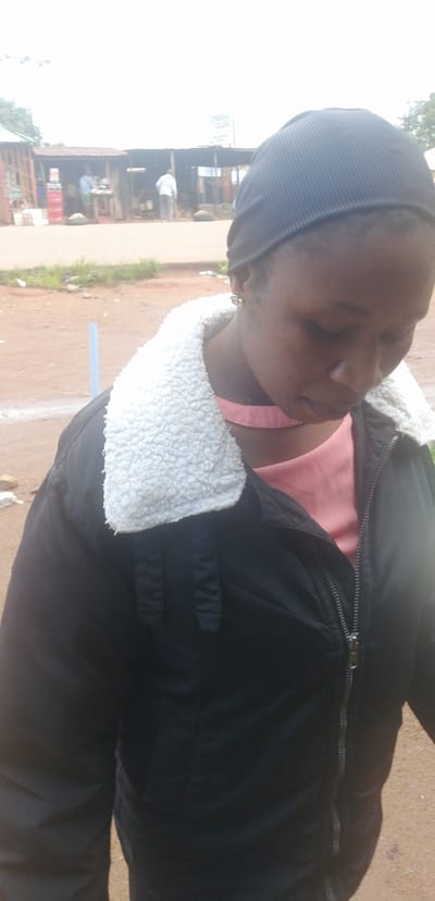 This media file shows a medium shot of a young Black woman wearing a black beanie and a black jacket with a white fuzzy collar. She is looking down and to the right, her face partially obscured by her downward gaze. She wears a pale pink, sleeveless top underneath her jacket. The background is somewhat out of focus, but appears to be an outdoor market scene, possibly Kassa Market in Nigeria, based on the provided location context.

In the background, there are structures that resemble market stalls, with corrugated metal roofs and open fronts. Several people are visible in the distance, appearing to be engaged in market activities. One person in a white shirt is standing near a stall. Another person, wearing a blue shirt, is partially visible on the left side. The ground is reddish-brown, indicative of dirt or unpaved road common in such locations. The sky is overcast and bright, suggesting it might be daytime with cloudy weather. There is a blue pole-like object visible in the mid-left background. The overall impression is of a bustling, everyday market environment. The woman's expression is difficult to discern due to her gaze, but she appears to be in a casual setting. No prominent text is visible in the image.