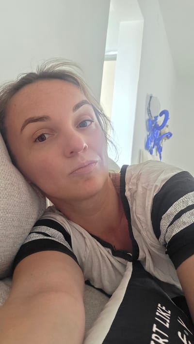 A woman is lying on her side on a couch, looking up at the camera. She is wearing a t-shirt with black and white stripes on the sleeves and text on the front. The text on her shirt is partially visible and reads "RT LIKE F Y". Her face is illuminated by a soft light, suggesting it is daytime. Her expression is neutral, and she appears relaxed.
In the background, a white ceiling is visible, with a window allowing natural light to enter. There is a bright blue object hanging from the ceiling, possibly a toy or decoration, with some text on it that is difficult to read. The scene appears to be indoors, in a residential setting. The lighting and the woman's relaxed posture suggest a casual and comfortable environment.
The image is oriented in a portrait orientation, with the woman's head towards the top right of the frame. The overall mood of the image is calm and intimate.