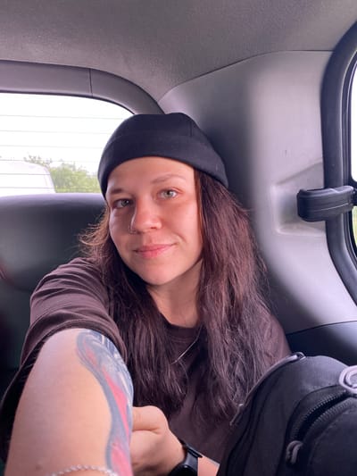 A young woman wearing a black beanie and a brown t-shirt is sitting in the back seat of a car. She has long, dark, wavy hair and a nose ring. Her left arm, visible in the foreground, is tattooed with abstract designs in shades of blue, pink, and grey. She is looking towards the camera with a slight smile. The interior of the car is visible, with a dark leather seat and the car's ceiling. The background shows a blurred view of greenery through the car window, suggesting an outdoor setting, possibly a road leading to or from Petrovskoye, Russia.